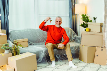 Older man sitting on his sofa, surrounded by moving boxes, smiling as he holds up the keys to his new home, symbolizing a successful move and new beginnings.