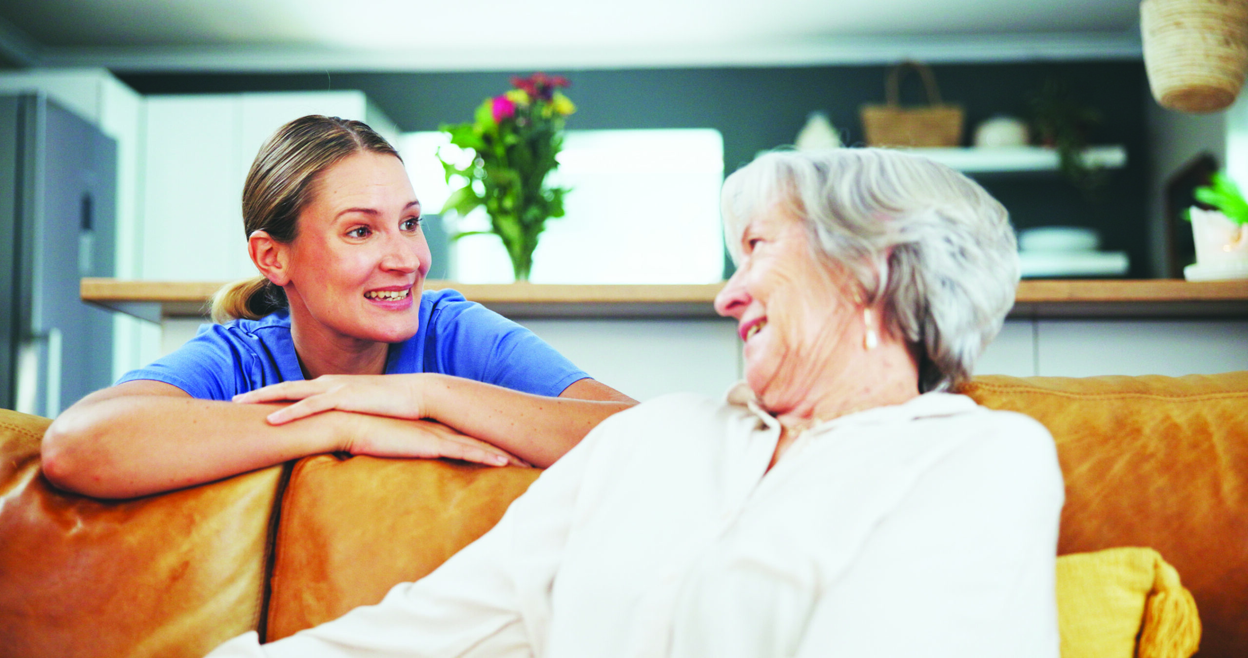 A hospice caregiver kneeling while speaking with an elderly woman who is seated, both smiling.