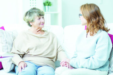 A hospice volunteer and a senior woman sitting together on a couch, holding hands and smiling.