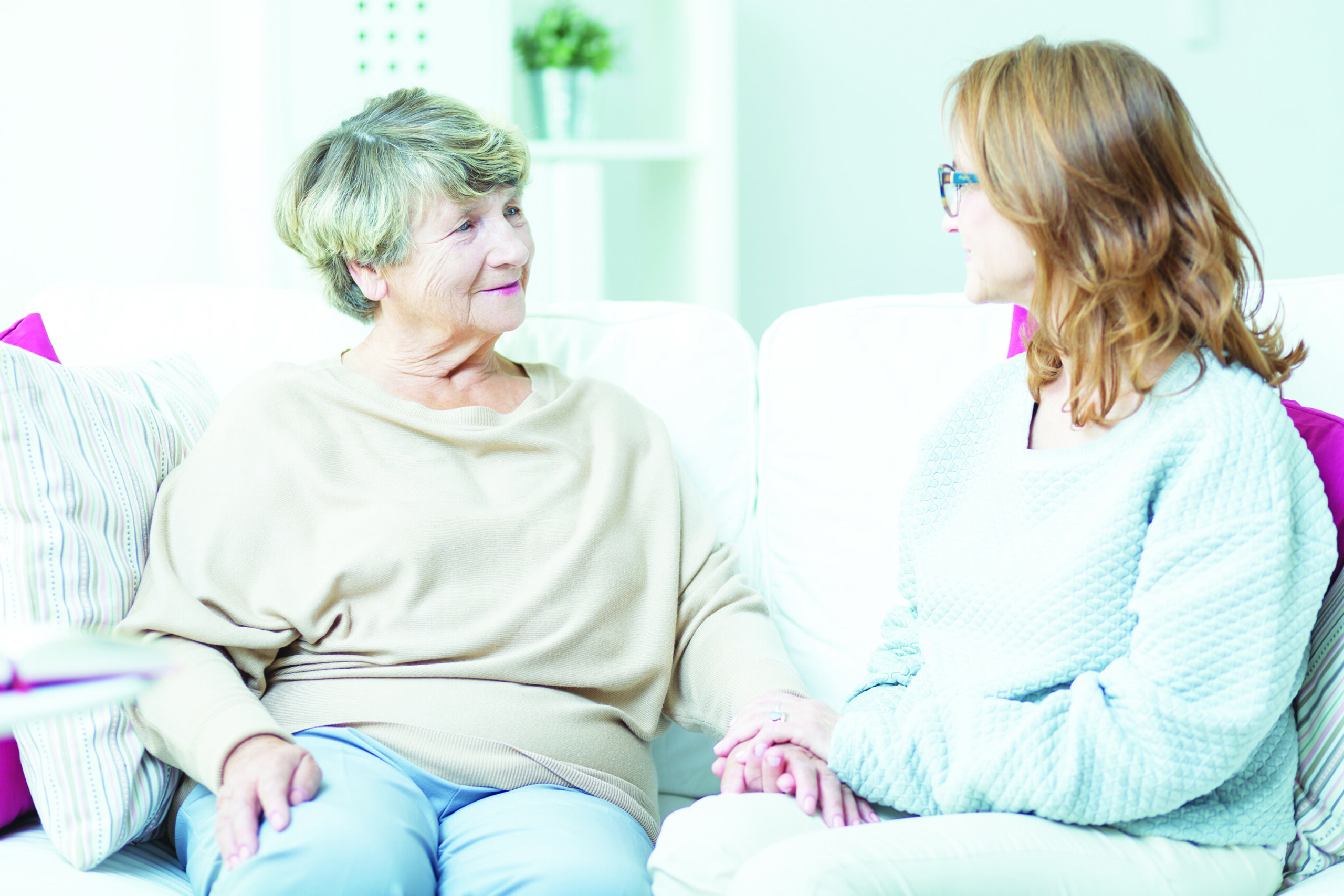 A hospice volunteer and a senior woman sitting together on a couch, holding hands and smiling.